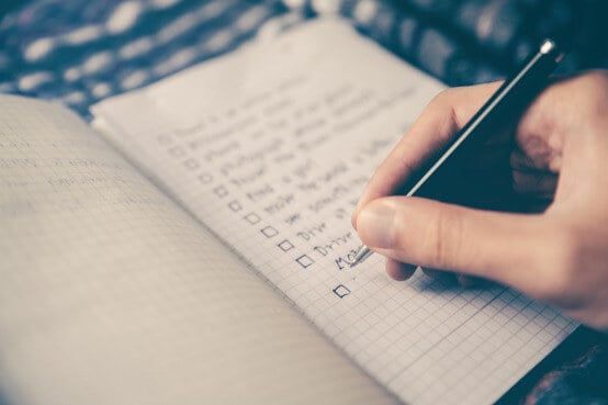 A hand writes with a pen on a checkered notebook page with checkboxes.